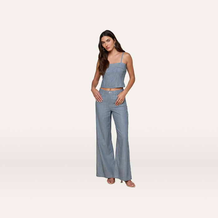 A woman stands against a plain background, wearing the Arlin Tank—a sleeveless blue stripe corset-style top—paired with matching high-waisted, wide-leg pants. Her long hair is down and she looks at the camera with her hands in her pockets.