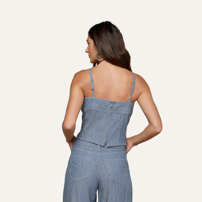 Woman facing away, wearing the Arlin Tank—a blue pinstriped sleeveless top with thin straps, a visible back zipper, and subtle corset-inspired detailing—paired with matching pinstriped pants. Her hands are in her pockets and she has long brown hair.