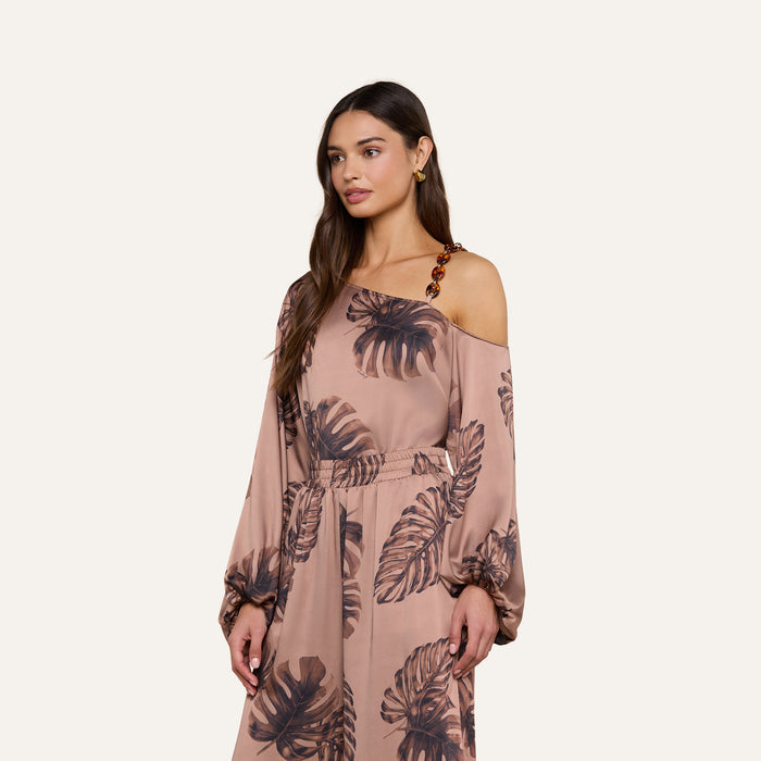 A woman models the Asami Chain One-Shoulder Blouse, a light brown top with dark leaf prints, long sleeves, an elastic waist, and a signature tortoise shell-inspired decorative strap, set against a plain light background.
