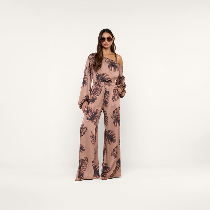 A woman wearing sunglasses and the Asami Chain One-Shoulder Blouse, featuring a mauve hue with a dark leaf pattern, stands confidently against a plain light background with one hand in her pocket.