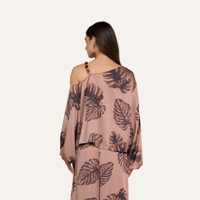 A woman with long brown hair models the Asami Chain One-Shoulder Blouse, a loose-fitting, light brown top with dark leaf prints. Featuring long sleeves and an elegant drape, she is pictured from behind against a plain background.