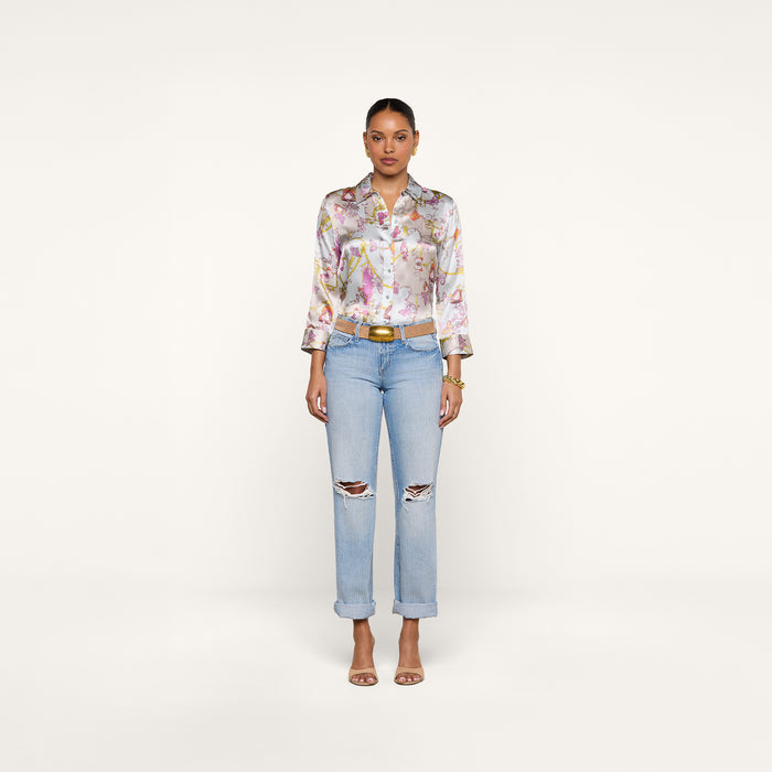 A woman stands facing forward against a plain white background, wearing a vintage-inspired floral button-up shirt, the Ashby Low-Rise Slouch Jean in light blue with ripped details, a belt, and heeled sandals.