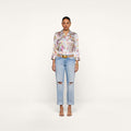 A woman stands facing forward against a plain white background, wearing a vintage-inspired floral button-up shirt, the Ashby Low-Rise Slouch Jean in light blue with ripped details, a belt, and heeled sandals.