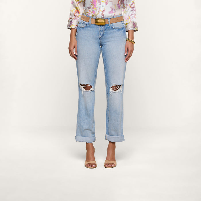 A woman models the Ashby Low-Rise Slouch Jean in light blue, featuring vintage-inspired rips and rolled cuffs. She pairs them with a floral blouse, brown belt, bracelets, and open-toe heels. Her hands are at her sides against a plain white background; her face is not visible.
