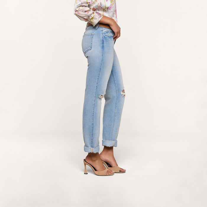 Wearing the Ashby Low-Rise Slouch Jean in vintage-inspired light blue with a small knee rip and cuffed hems, styled with a floral blouse and beige high-heeled sandals, the person stands sideways against a plain white background.