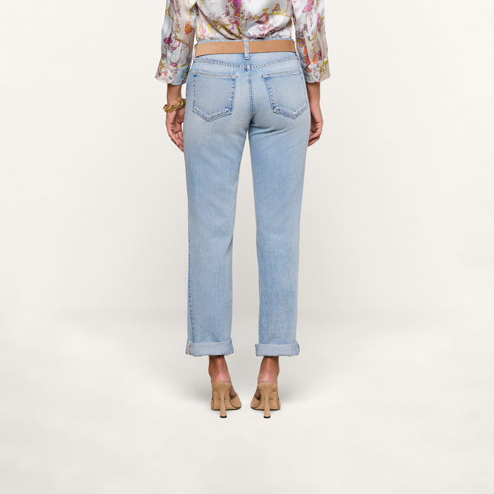A person stands facing away against a plain white background, wearing the Ashby Low-Rise Slouch Jean in a vintage-inspired light wash, paired with a floral blouse and beige heels.