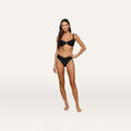 A woman stands barefoot against a white background, wearing the Ashleigh Bikini Bottom in black with V-cut and trapunto stitching. She has long brown hair, one hand resting on her hip, and a neutral expression.