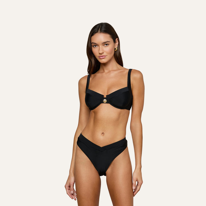 Woman wearing the Ashleigh Bikini Bottom, a black V-cut bikini with Trapunto stitching, paired with an underwire top featuring a central gold button, standing against a plain off-white background.