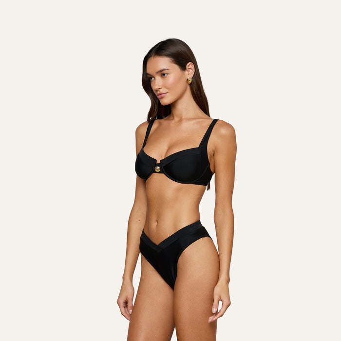 A woman stands against a plain background wearing the Ashleigh Bikini Bottom in black, styled with high-waist bottoms and a structured V-cut top featuring Trapunto stitching and a central gold accent.