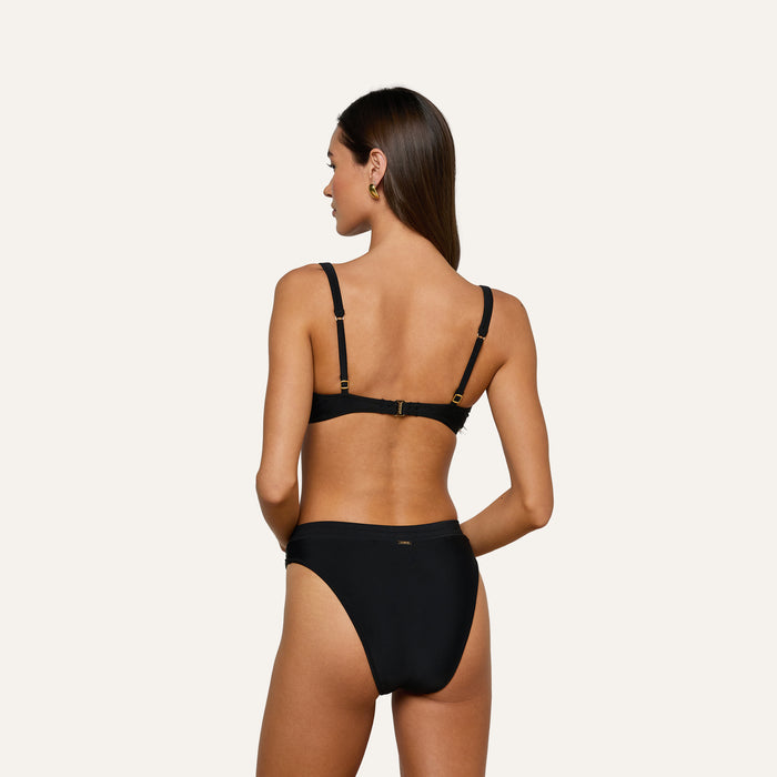 Woman with long brown hair stands with her back to the camera, wearing the Ashleigh Bikini Bottom, featuring black fabric, gold hardware, and Trapunto stitching, against a plain light background.