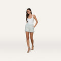 A woman stands with her hands in the pockets of the Ashley Tweed Short—chevron-patterned shorts with fringed trim—paired with a white tank top and beige high-heeled sandals against a plain white background.
