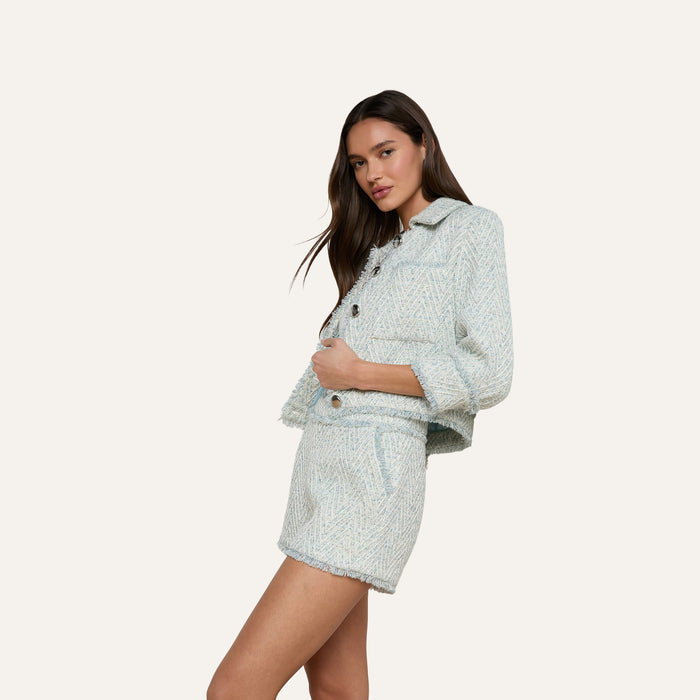 A woman poses in the Ashley Tweed Short—a pair of chevron-patterned, light blue tweed shorts—paired with a coordinating fringed jacket, showcasing a classic silhouette against a plain, off-white background.