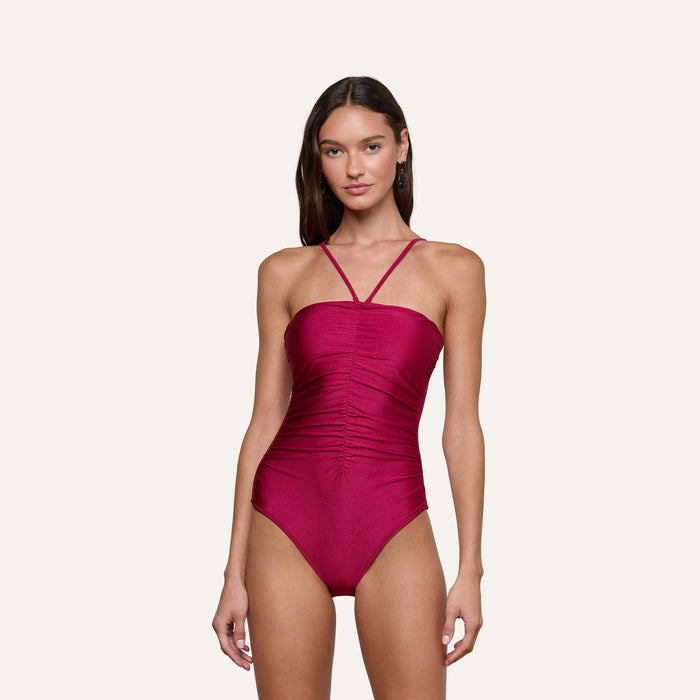 A woman stands wearing the Aubrey Ruched One-Piece Swimsuit, a sleeveless burgundy design featuring a high-cut leg, thin straps, and textured shirring. The background is plain and light-colored.