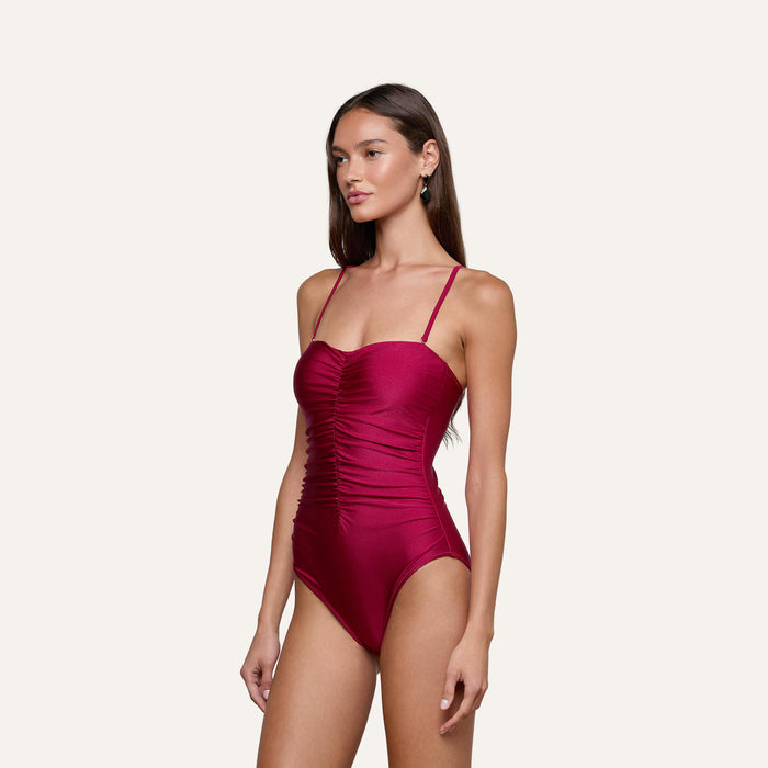 A woman wears the Aubrey Ruched One-Piece Swimsuit, featuring a dark red color, textured shirring, and thin shoulder straps, while standing against a plain light background.