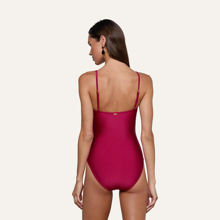 A woman with long brown hair stands with her back to the camera, modeling the Aubrey Ruched One-Piece Swimsuit in red. The swimsuit features adjustable straps and textured shirring. She wears black earrings, and the background is plain and light-colored.