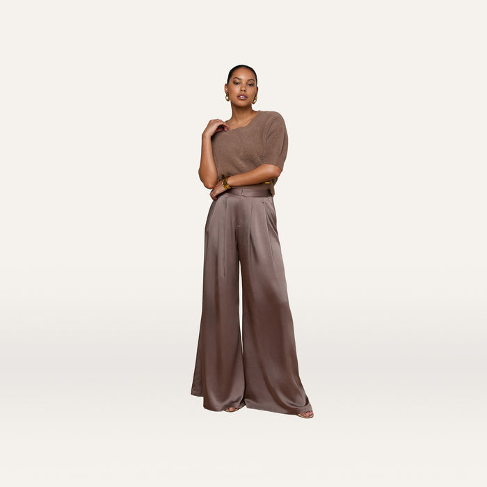 A woman stands against an off-white background, wearing a cardamom brown short-sleeve knit top paired with the Aubrie Satin Pant—a high-waisted, wide-leg trouser in a matching shade—styled with large earrings and open-toe shoes.