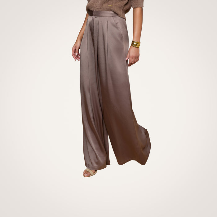Wearing the Aubrie Satin Pant in taupe, a person pairs it with a cardamom brown knit top, gold bracelets, and sandals while posing against a plain light background.