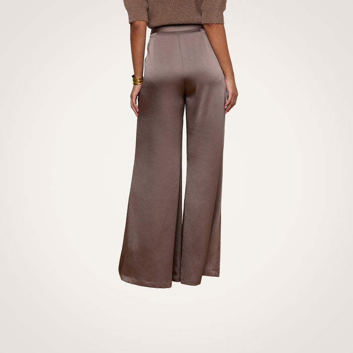 A person stands facing away in the Aubrie Satin Pant, featuring a high-waisted, wide-leg silhouette in taupe, paired with a cardamon brown sweater. The plain light background emphasizes the back of the trousers.