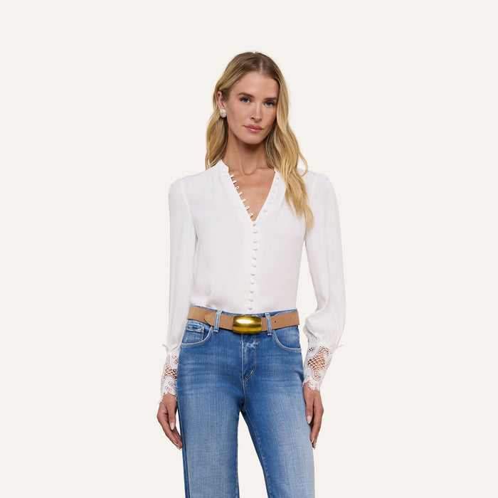 A woman with long blonde hair wears the Ava Blouse, a vintage-inspired ivory top featuring scalloped guipure lace cuffs, paired with blue jeans. She stands facing forward against a plain light background.
