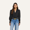 Woman with long brown hair wearing the Ava Blouse, a vintage-inspired black top with guipure lace cuffs, paired with blue jeans and standing against a plain light background.