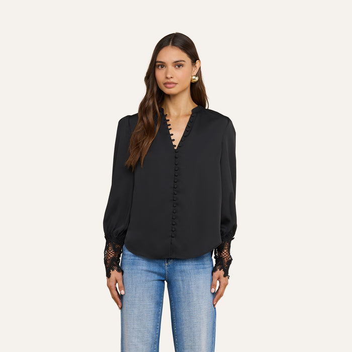A woman wearing the Ava Blouse, a vintage-inspired black top featuring button details and guipure lace cuffs, paired with light blue jeans and standing against an off-white background.