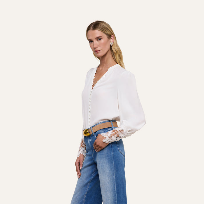 A woman poses against a plain, light background wearing the Ava Blouse—an ivory, vintage-inspired top featuring scalloped guipure lace cuffs—paired with blue high-waisted jeans.