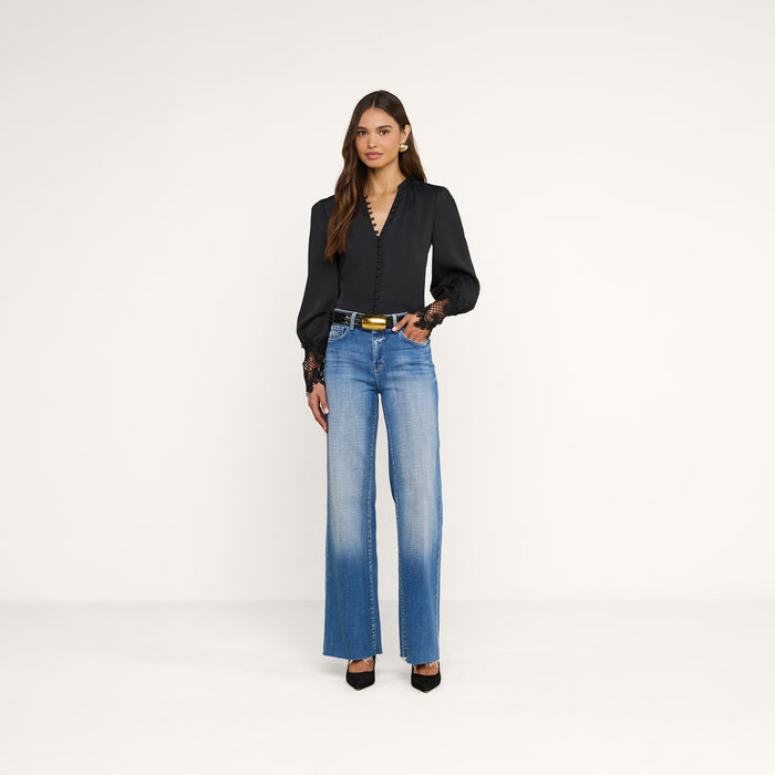 A woman stands against a plain white background wearing the Ava Blouse, styled with blue high-waisted jeans, a black belt featuring a gold buckle, and black heels.