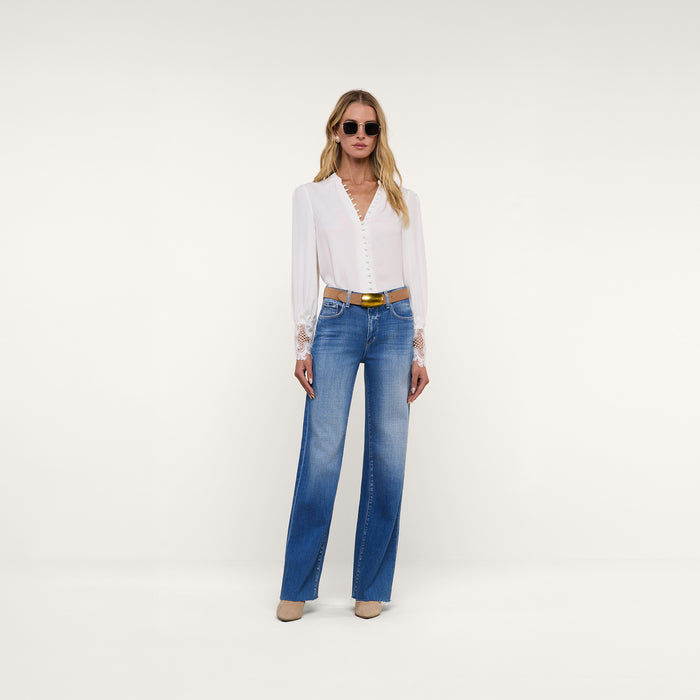 A woman wearing sunglasses, the Ava Blouse—a vintage-inspired ivory top with scalloped guipure lace cuffs—blue high-waisted jeans, a black belt, and tan shoes stands confidently against a plain white background.