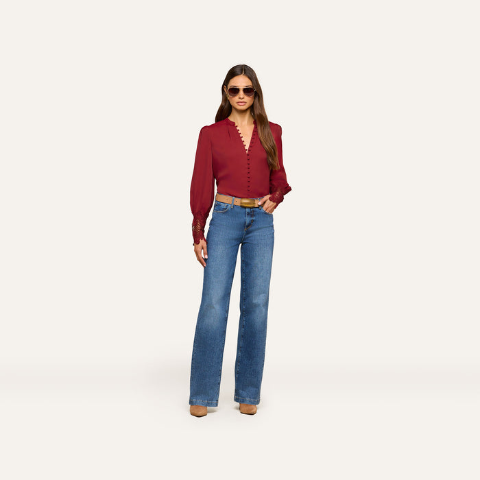A person wearing the Ava Blouse—a vintage-inspired burgundy top with puff sleeves—paired with blue high-waisted jeans, brown heeled shoes, and sunglasses stands against a plain light background.