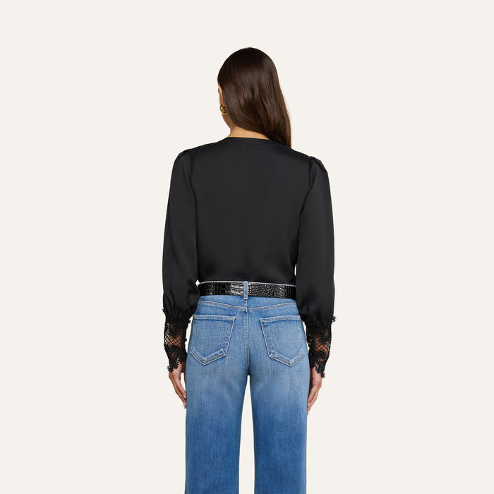 A woman with long brown hair stands facing away, wearing the vintage-inspired Ava Blouse—black with lace cuffs—and blue jeans. Her arms rest at her sides against a plain light background.