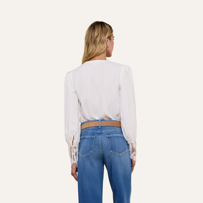 A woman stands facing away, wearing the Ava Blouse—a vintage-inspired ivory top featuring scalloped guipure lace cuffs—paired with blue jeans. Her blonde hair is worn down, and the background is plain and light-colored.