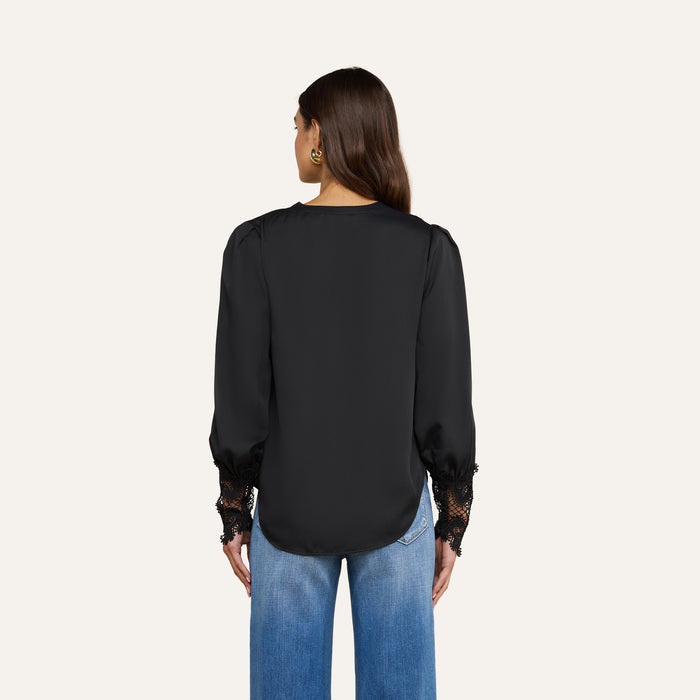 A woman with long brown hair, wearing the Ava Blouse—a vintage-inspired black top with lace cuffs—and blue jeans, stands facing away from the camera against a plain light background.