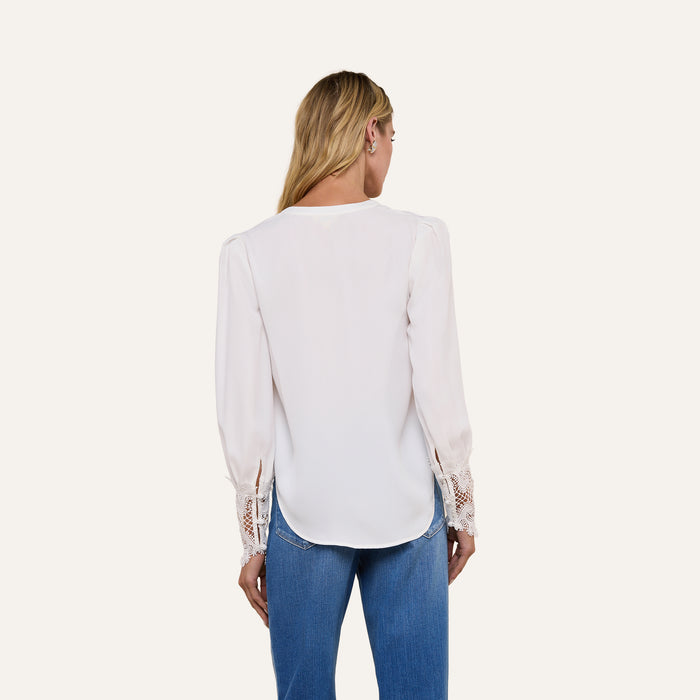A woman with long blonde hair is seen from behind, wearing the vintage-inspired Ava Blouse in ivory with scalloped guipure lace cuffs, paired with blue jeans, against a plain light background.