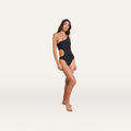 A woman stands barefoot against a plain white background, wearing the Ava Chain One-Piece Swimsuit—a black, one-shoulder swimsuit featuring a curve-sculpting cutout and removable chain detail on the right side.