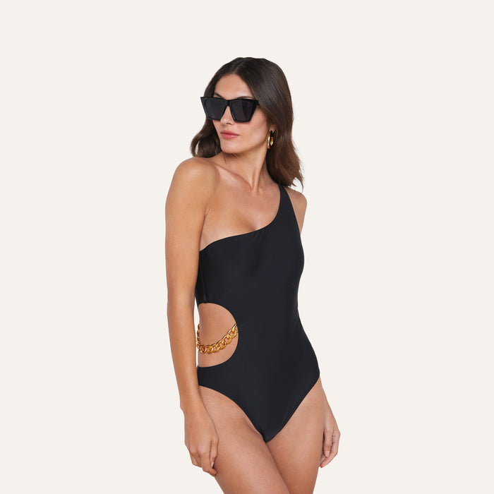 A woman models the Ava Chain One-Piece Swimsuit—a black, curve-sculpting one-shoulder design featuring a gold chain detail at the waist—paired with sunglasses and gold hoop earrings, standing against a plain light background.