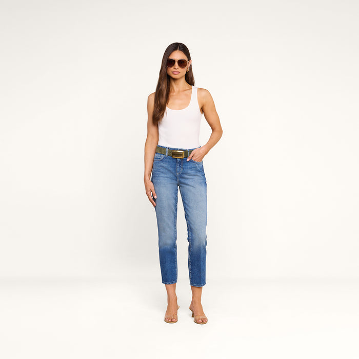 A woman wearing sunglasses, a white tank top, the Avena Slim-Leg Jean, and heeled sandals stands against a plain white background with her hands in her pockets.