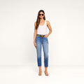 A woman wearing sunglasses, a white tank top, the Avena Slim-Leg Jean, and heeled sandals stands against a plain white background with her hands in her pockets.