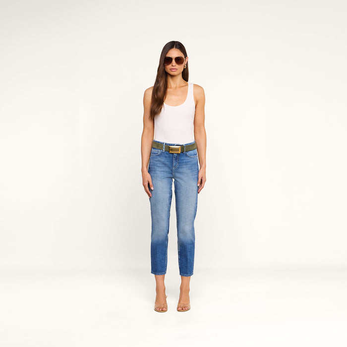 A woman stands against a plain white background, facing forward with her arms relaxed at her sides, wearing sunglasses, a white tank top, clear heels, and the Avena Slim-Leg Jean with a belt.