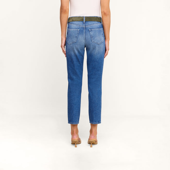 A person stands with their back to the camera, wearing a white sleeveless top, beige heels, and the Avena Slim-Leg Jean in a blue high-rise slim-cut denim style against a plain light background.