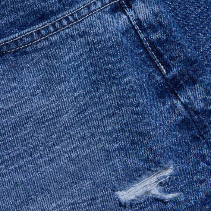 Close-up view of blue destructed denim fabric showing a seam and a small ripped, frayed hole, highlighting the texture and worn appearance of the Zelda Slouchy Straight Jean.
