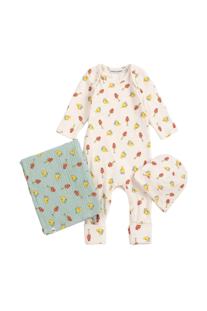 A baby clothing set laid flat on a light gray background, including a long-sleeve footless one-piece in warm off-white with a repeating yellow and red mushroom print, a folded pale blue quilted blanket with the same mushroom pattern, and a matching soft hat.