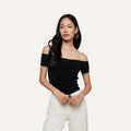 A woman with long dark hair wears the black Baliegh Ruched Bodysuit—an off-the-shoulder rayon stretch jersey top—paired with white pants, a white belt, and hoop earrings, standing against a plain light background.