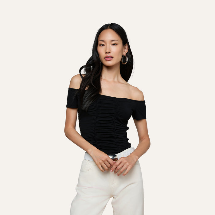 A woman with long dark hair wears the black Baliegh Ruched Bodysuit—an off-the-shoulder rayon stretch jersey top—paired with white pants, a white belt, and hoop earrings, standing against a plain light background.
