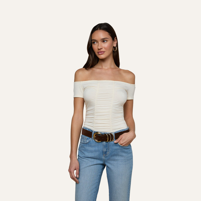 A woman wearing the Baliegh Ruched Bodysuit in vintage white, paired with blue jeans and a brown belt, stands with one hand in her pocket against a plain background.