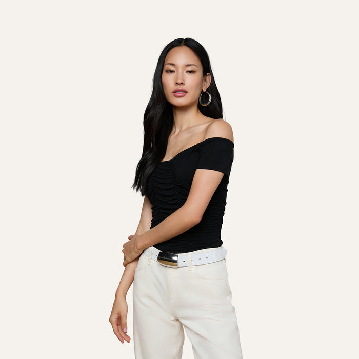 A woman with long dark hair stands against a plain light background, wearing the Baliegh Ruched Bodysuit—a black off-the-shoulder rayon stretch jersey top—paired with large hoop earrings and white high-waisted belted pants.