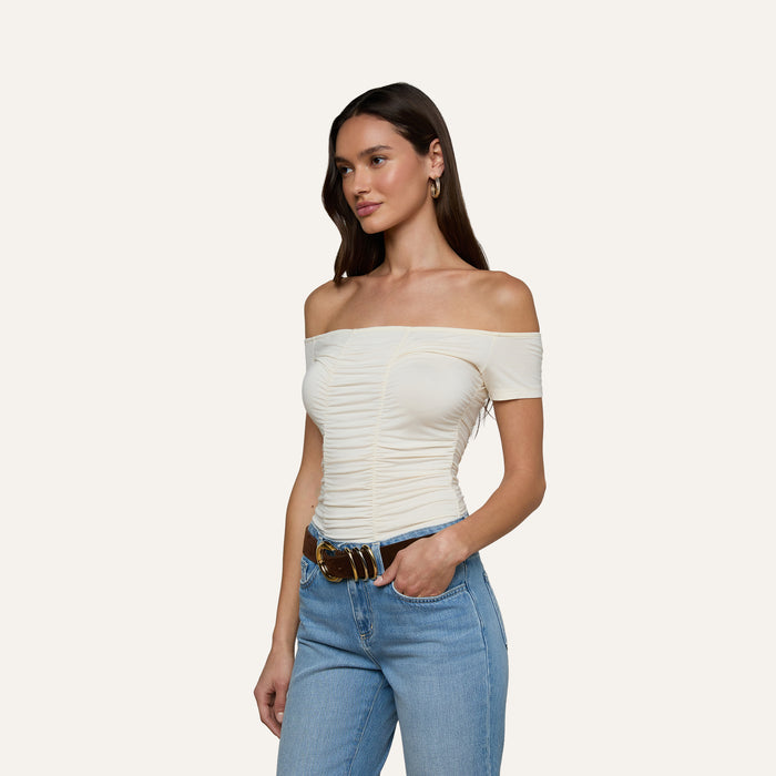Woman wearing the Baliegh Ruched Bodysuit in vintage white, paired with light blue jeans, a brown belt, and gold hoop earrings, standing against a plain light background.