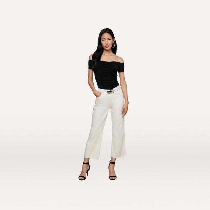 Woman wearing the Baliegh Ruched Bodysuit with an off-the-shoulder neckline, paired with white wide-leg pants, a black belt, and black high-heeled sandals, standing against a plain light background with her hands in her pockets.