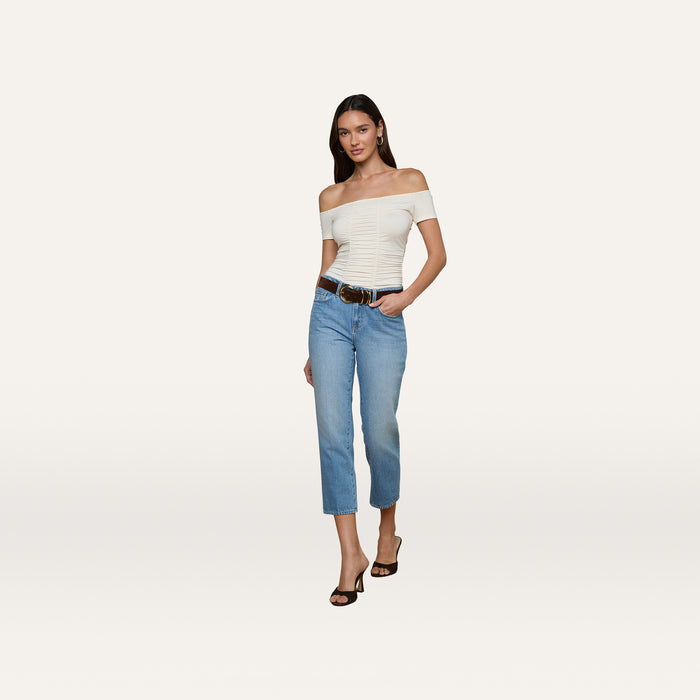 A woman wearing the Baliegh Ruched Bodysuit—an off-the-shoulder vintage white top—paired with light blue jeans, a black belt, and black heels stands confidently against a plain white background.