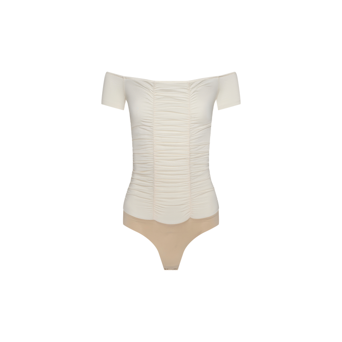 The Baliegh Ruched Bodysuit is an off-the-shoulder, vintage-inspired white top with short sleeves, delicate scalloped edges, and flattering ruching. It features a beige thong bottom and is displayed on a plain white background.