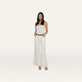 A woman stands against a plain white background wearing a white tank top, the Beau Wide-Leg Jean in high rise stretch denim, a black belt, and beige heels, with her hands in her pockets.